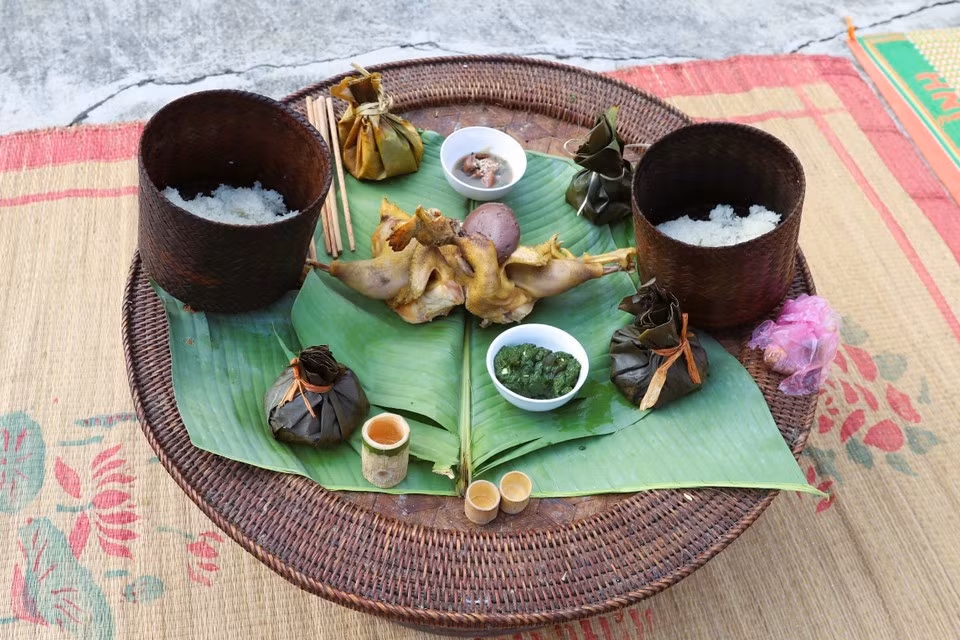 Las ofrendas sobre la bandeja ceremonial incluyen pollo hervido, arroz glutinoso, licor servido en una copa de bambú, arroz con pescado o carne y especias envueltos en hoja de plátano, y ensalada de musgo de arroyo servida en cuencos de porcelana.
